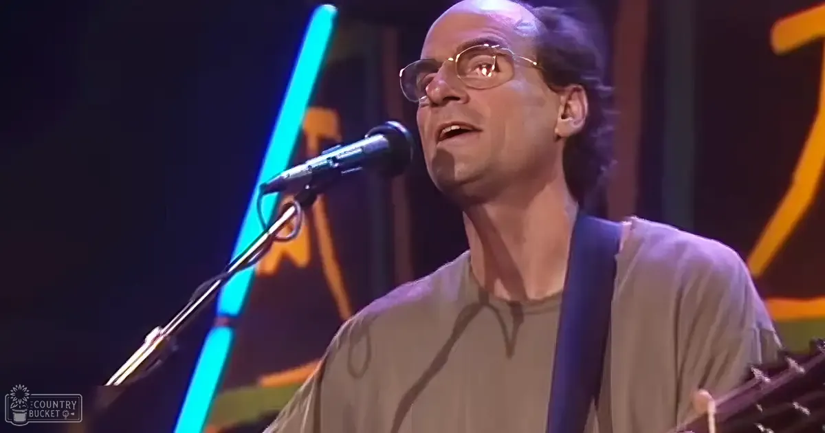 A middle-aged man in a relaxed fit, crew neck t-shirt with thin-framed glasses performs on stage at an indoor concert venue under artificial blue neon and orange lights.