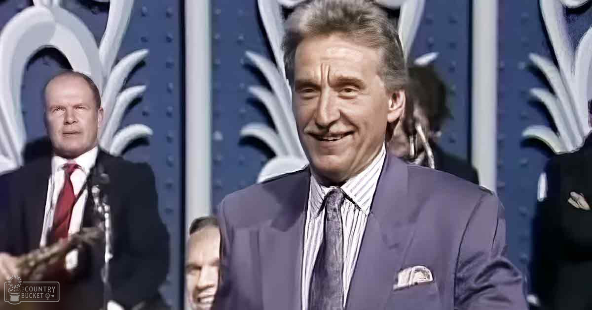 A middle-aged man in a dark suit jacket and light-colored shirt with a patterned tie stands at an event or ceremony, alongside another adult male wearing similar attire but with a red tie. The setting includes decorative paneling with white ornate designs on a blue backdrop.