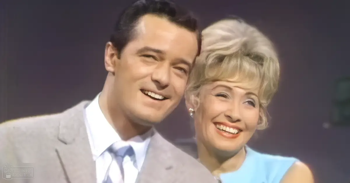 An adult male singer in a mid-20th-century gray suit jacket with a white shirt and blue striped tie, accompanied by an adult female wearing a light blue sleeveless top. The scene appears to be set indoors, possibly on stage or in a studio environment.