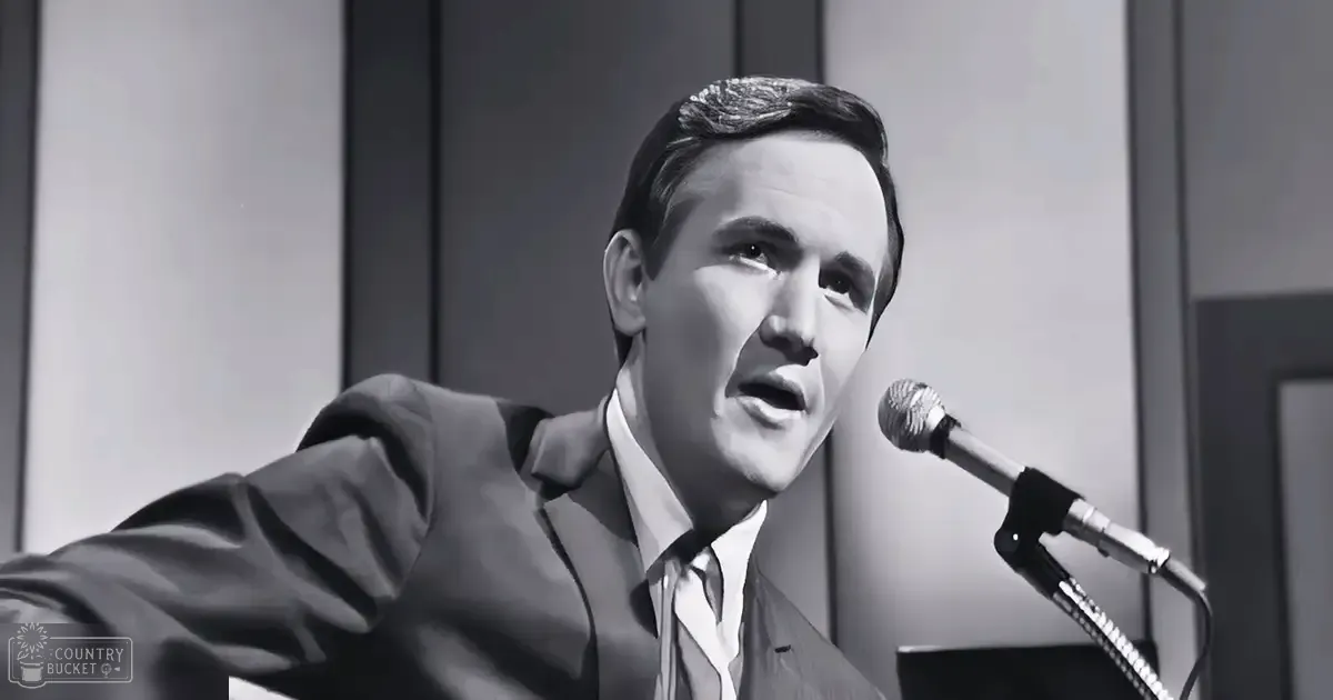 Adult male in a dark suit jacket with a white collar shirt against an indoor backdrop of vertical paneling or curtains, likely a recording studio or performance space.