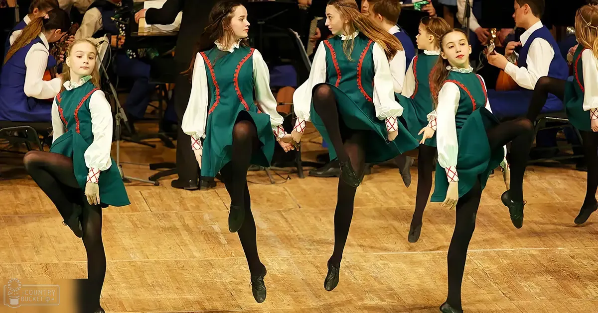 A group of young female children performing Irish step dancing on a stage with wooden flooring, under artificial lighting.