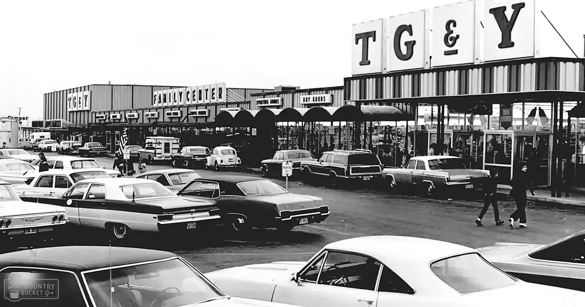 An outdoor shopping center with multiple stores, typical of a mid-20th-century urban environment; spring or summer setting, daytime light, black and white tones from the grayscale image.