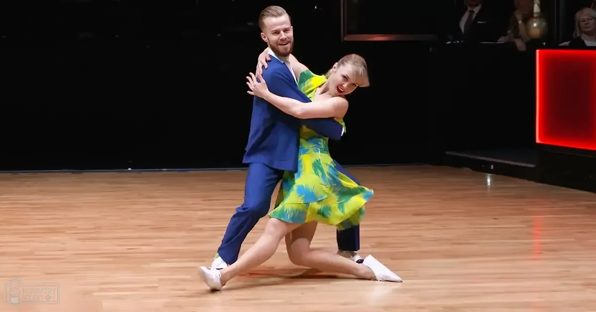 A couple dancing in an indoor dance hall with a polished wooden floor and bright lighting, wearing a blue suit and white sneakers for the man and a yellow dress with green floral patterns for the woman.