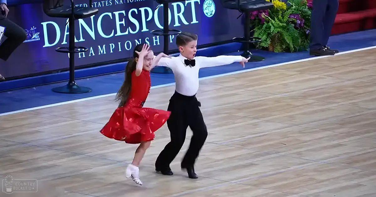 A young couple dancing at an event called 'United States National Dancesport Championship'. The girl wears a red, shiny dress with white ballet slippers. The boy is in a white shirt, black pants, and a bow tie.