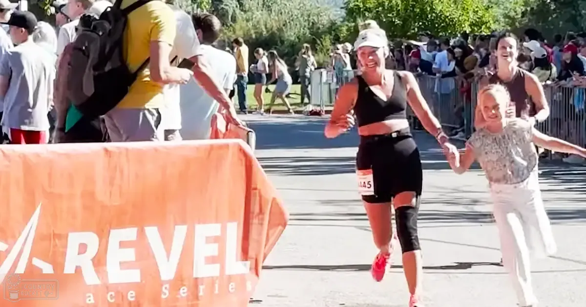 An adult female runner in a Revel Race Series event crossing the finish line, accompanied by a child in light-colored pants and a patterned top, surrounded by spectators and greenery under clear daytime skies with orange banners visible.