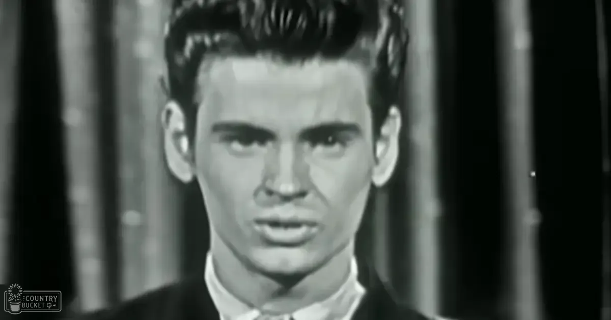 A young adult male wearing a dark suit jacket over a white collared shirt with a tie knot stands indoors against vertical blinds, suggesting a formal or professional setting.