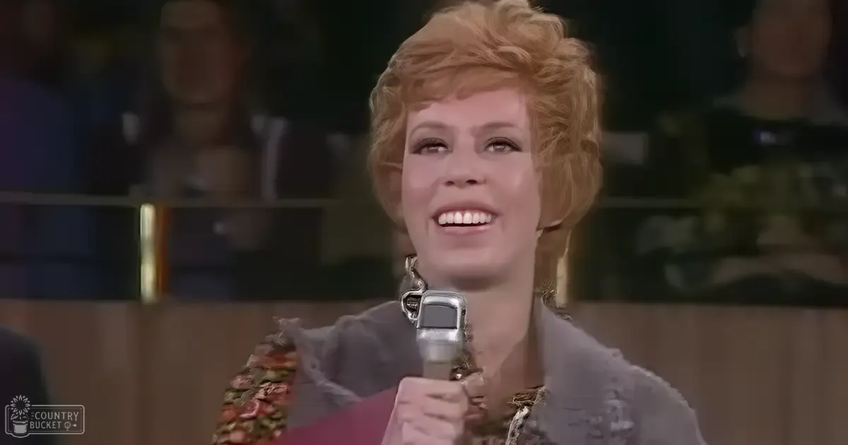 A middle-aged female performer in a stage setting, wearing a floral-patterned garment with textured overlays, holding a microphone.