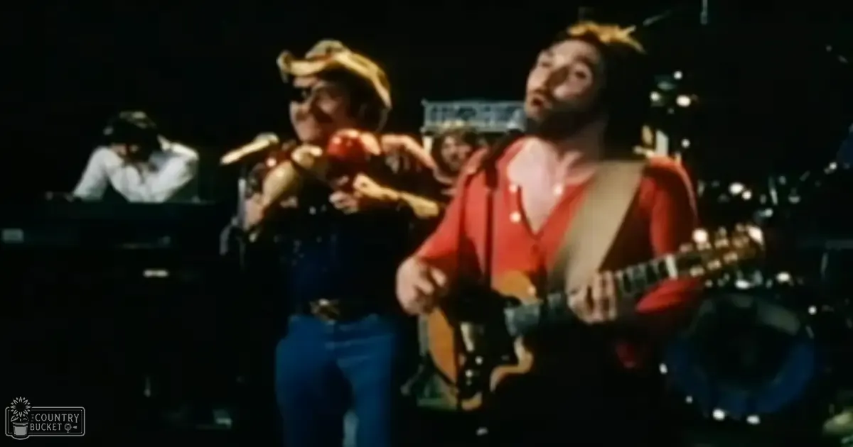 A musical performance scene with three adult male performers in a concert hall, wearing red jackets and blue jeans. The background features stage lighting and musical instruments.