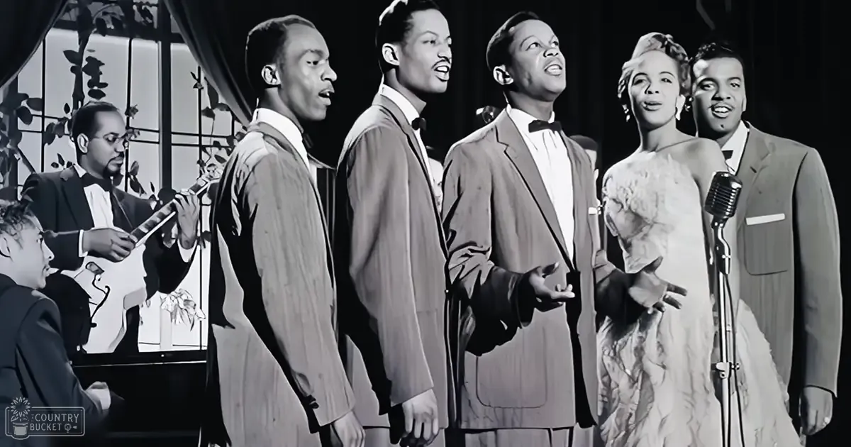 A musical performance with adult males in suits on a stage, indoor setting with curtains and stage lighting.