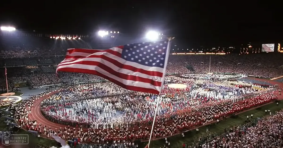 A nighttime Olympic stadium scene illuminated by bright floodlights, with a large American flag waving prominently in front of tiered rows filled with spectators dressed in white uniforms with red accents.