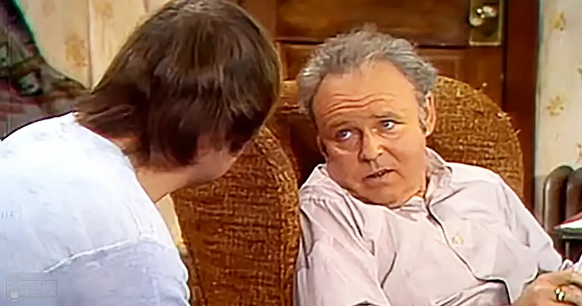 An older adult male seated on a wicker chair indoors, wearing a light-colored button-up shirt and a ring; another person stands nearby in a home setting with floral wallpaper.