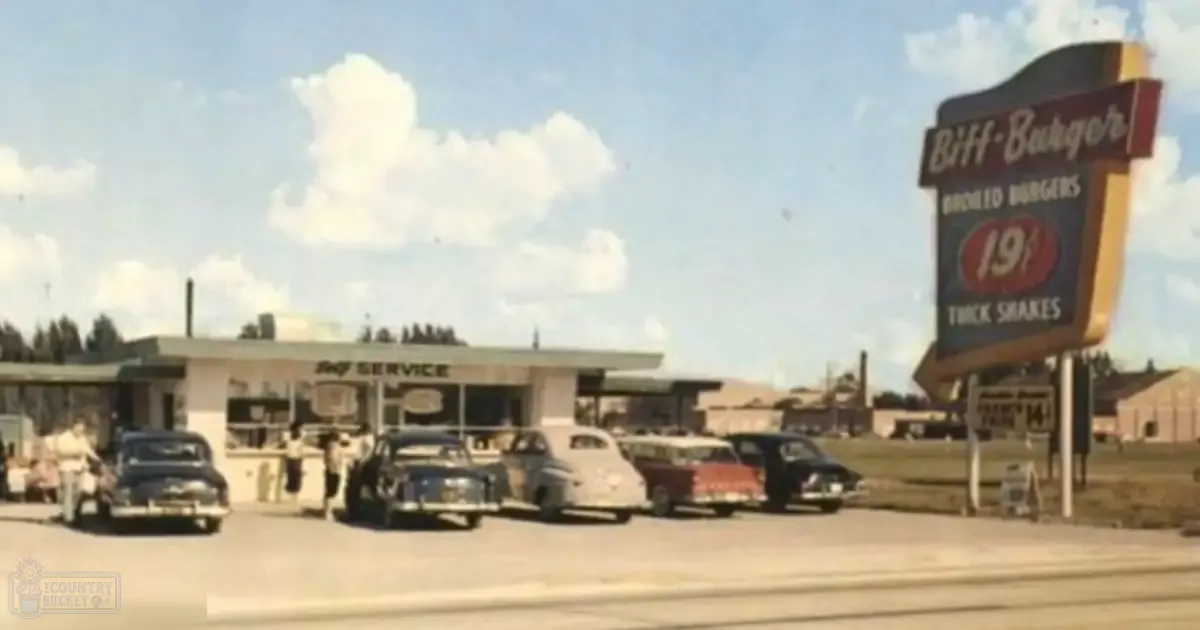 Adults at Biff-Burger drive-in restaurant with vintage cars and a large sign advertising broiled burgers in an open, rural setting under a clear sky.