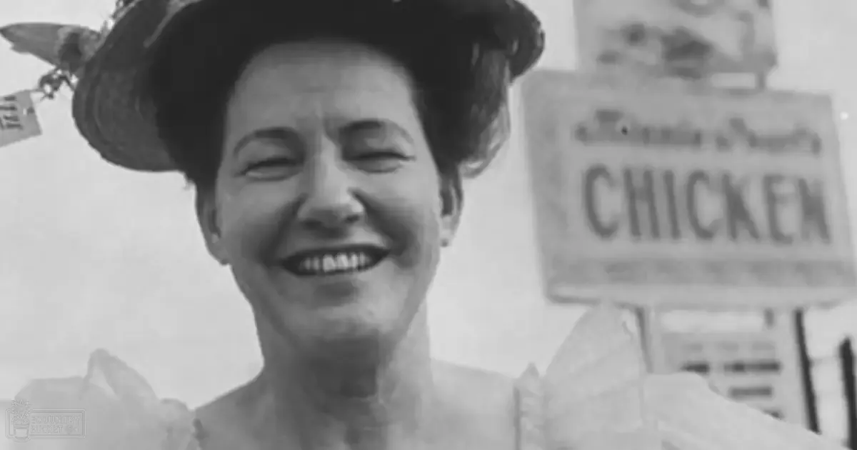 An older adult woman wearing a vintage-style light-colored garment with ruffled shoulders and neckline stands outdoors in a rural setting with 'CHICKEN' signage behind her.