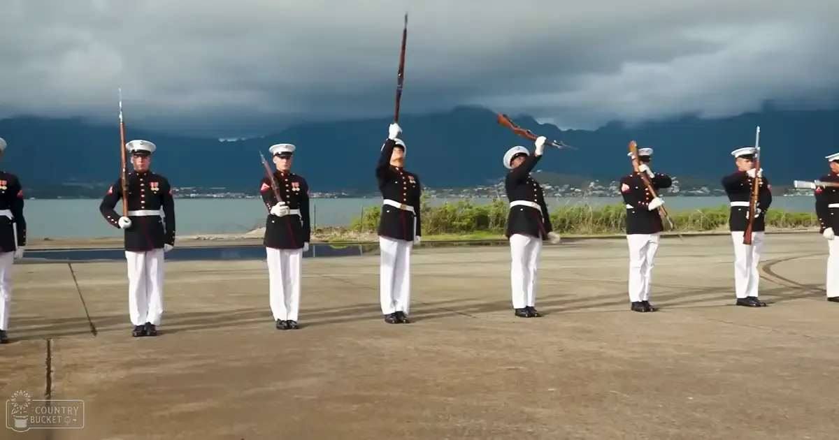 A group of young adult male soldiers in dark blue jackets with red trim, white trousers, black shoes, and white caps perform a ceremonial sword routine on an airfield near water with mountains under cloudy skies.