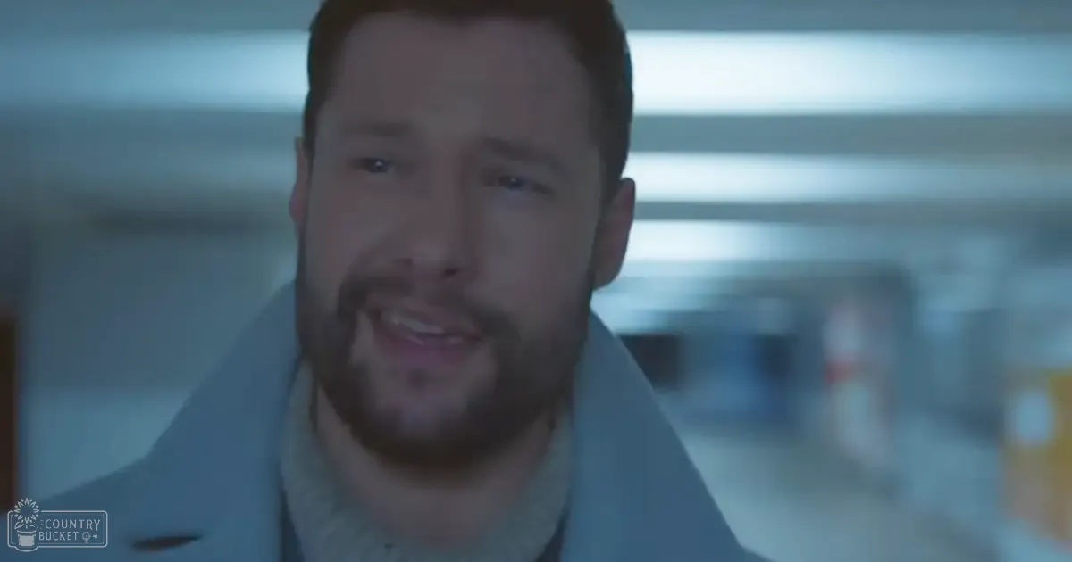 Middle-aged adult male in a relaxed casual light-gray jacket with high collar, seated indoors under artificial lighting, with blurred background suggesting an indoor public space like a hallway or corridor.