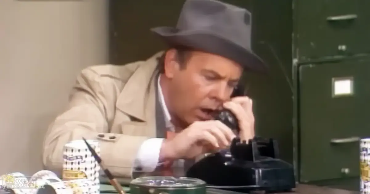 A middle-aged adult male in a tailored beige trench coat, speaking on an old-fashioned rotary phone in a professional office setting with green filing cabinets and a glass ashtray on the desk.