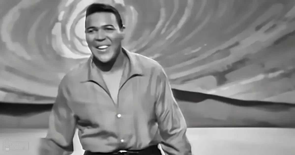 Young adult male wearing a light gray, relaxed fit long-sleeved button-up shirt with collar and pocket detail, indoors against a backdrop resembling clouds or abstract art.