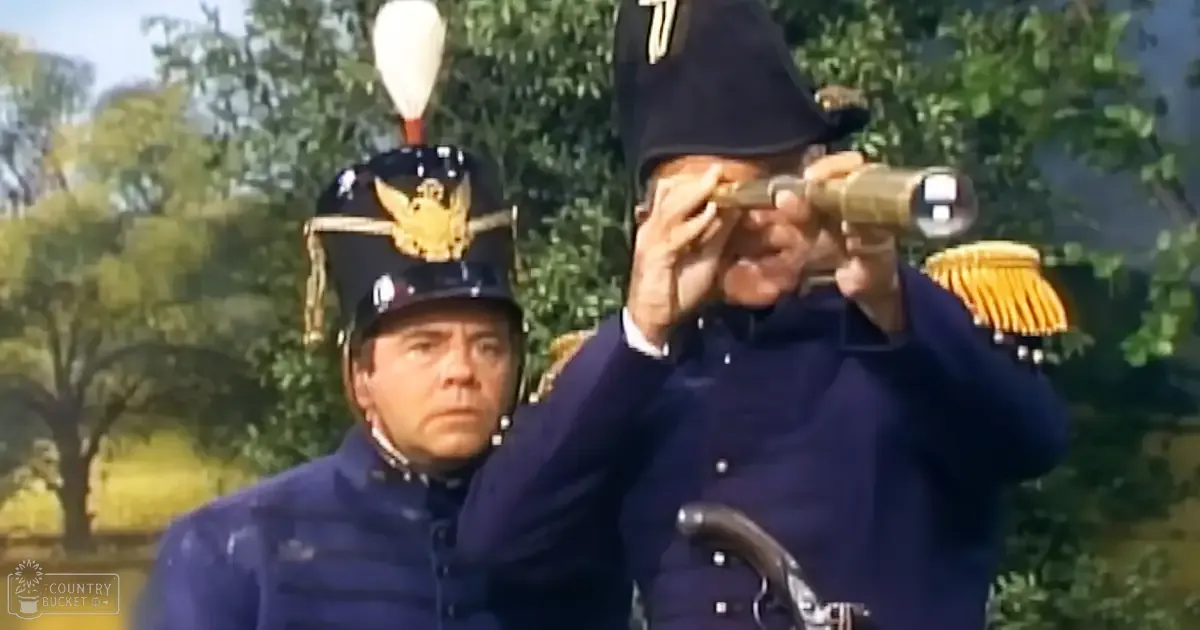 A historical drama scene set outdoors with lush greenery and clear skies. Adults wearing dark blue military uniforms are seated, one holding a telescope.