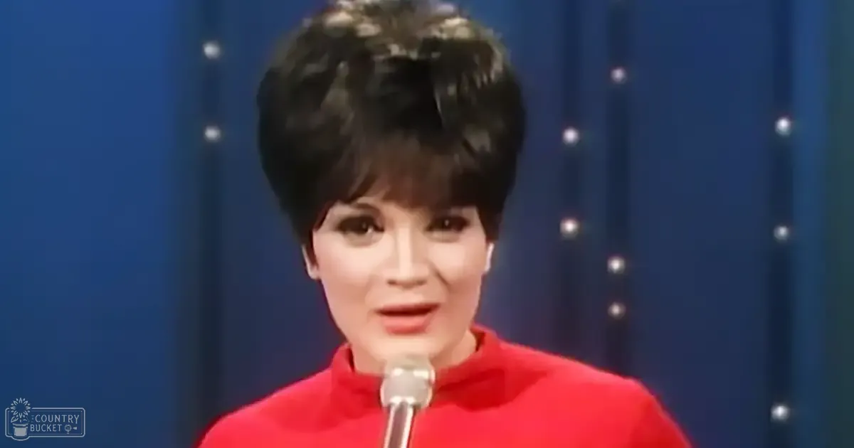An adult female in a red, fitted 1960s-style dress with short sleeves stands before a microphone and blue star-patterned curtain backdrop.