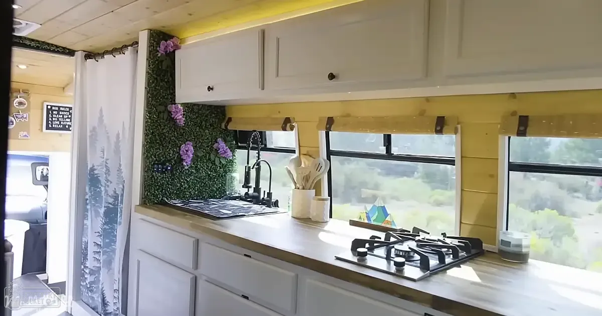 A kitchen interior in an RV or mobile home, with white cabinets with dark handles, a wooden countertop, a gas stove with four burners, and a sink with a modern faucet. The setting includes a green wall panel with purple flowers, a white mug holding kitchen utensils, and a small decorative geometric sculpture on the counter.