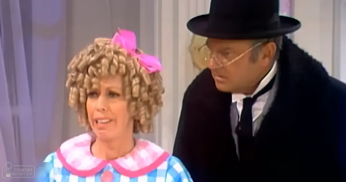 A middle-aged woman in a loose, playful blue and white checkered dress with pink trim on the collar stands beside an older adult man wearing a dark blue suit jacket over a white shirt and bow tie. They appear to be part of a performance or skit against a simple curtain backdrop.