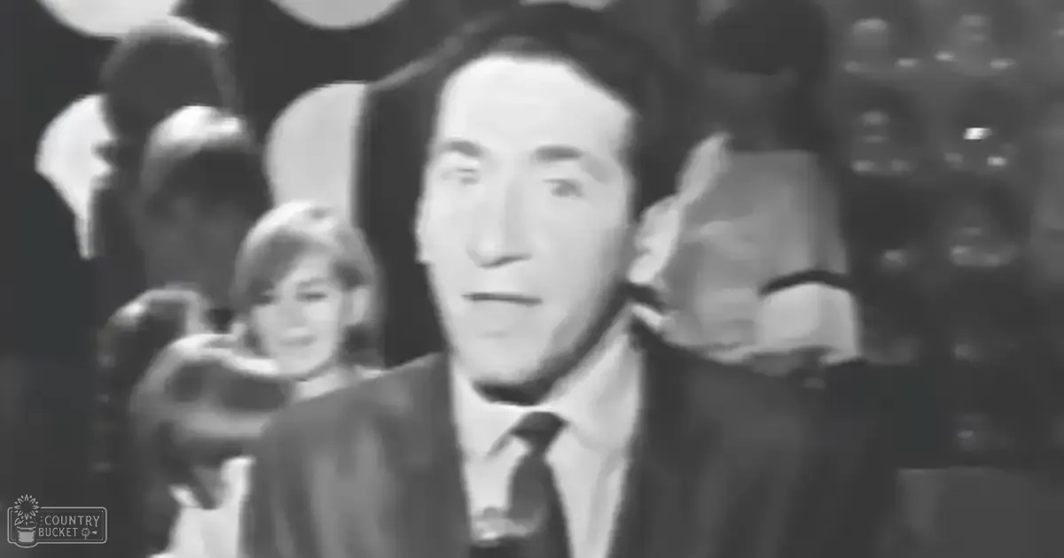 Adult male wearing a dark suit with tie at an indoor formal event; young adult female behind him in light-colored attire, likely from mid-20th century style.