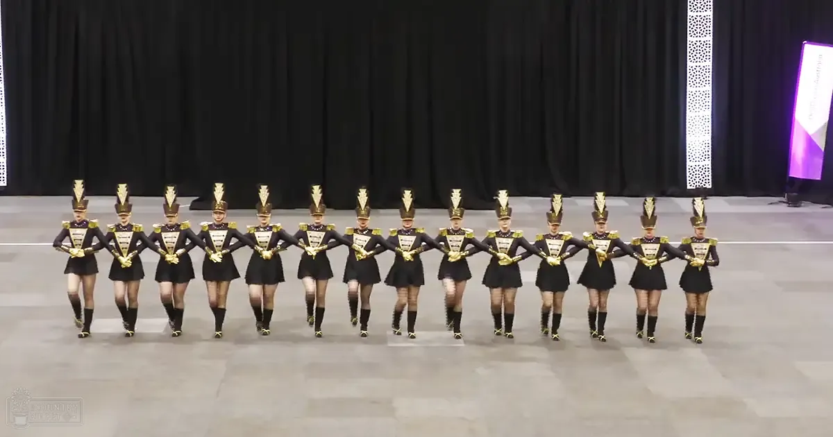 A marching band performance in an indoor arena, with young adult female performers in form-fitting black and gold uniforms with tall hats and high-heeled boots. The setting includes a stage floor and black curtains under artificial lighting.