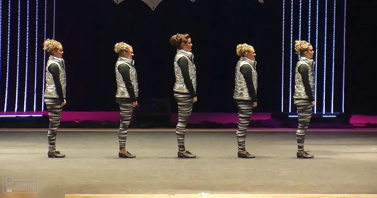 A group of young adult female performers on stage, wearing silver reflective vests and striped leggings, in a theater or dance hall setting with purple lighting.
