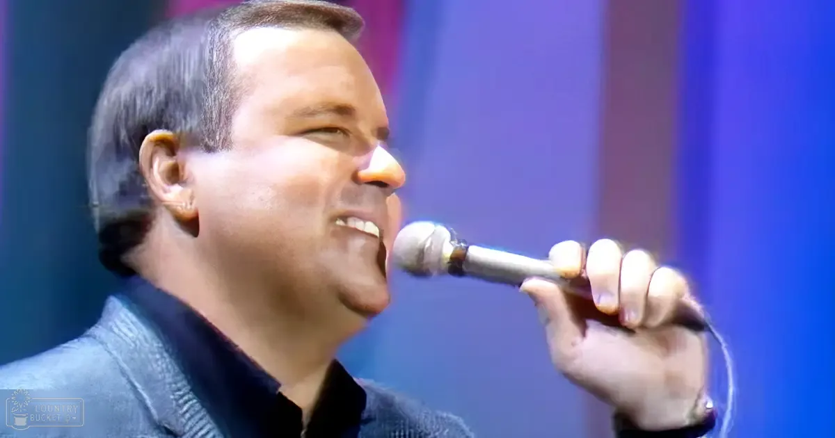 A middle-aged man wearing a dark textured jacket stands against a stage setting under blue lighting during what appears to be a concert or speaking event.