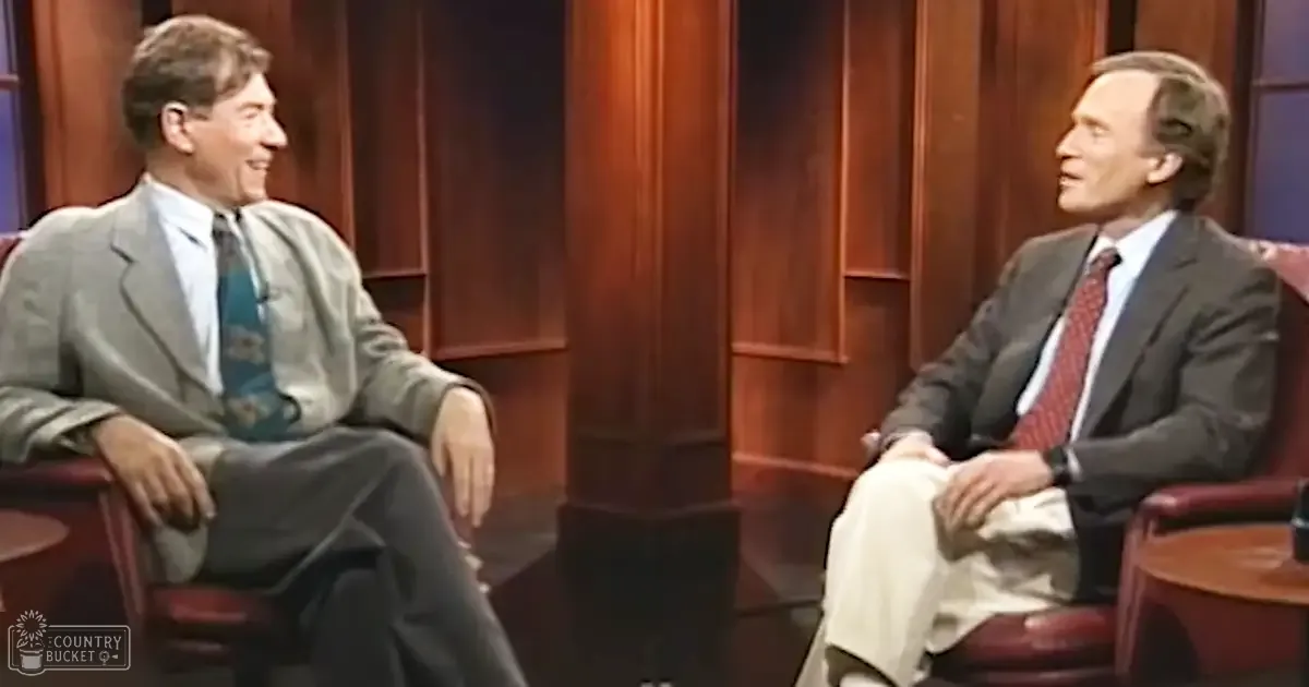 Two adult men in a studio setting, wearing formal suits from the late 20th century with red leather armchairs and dark wood paneling.