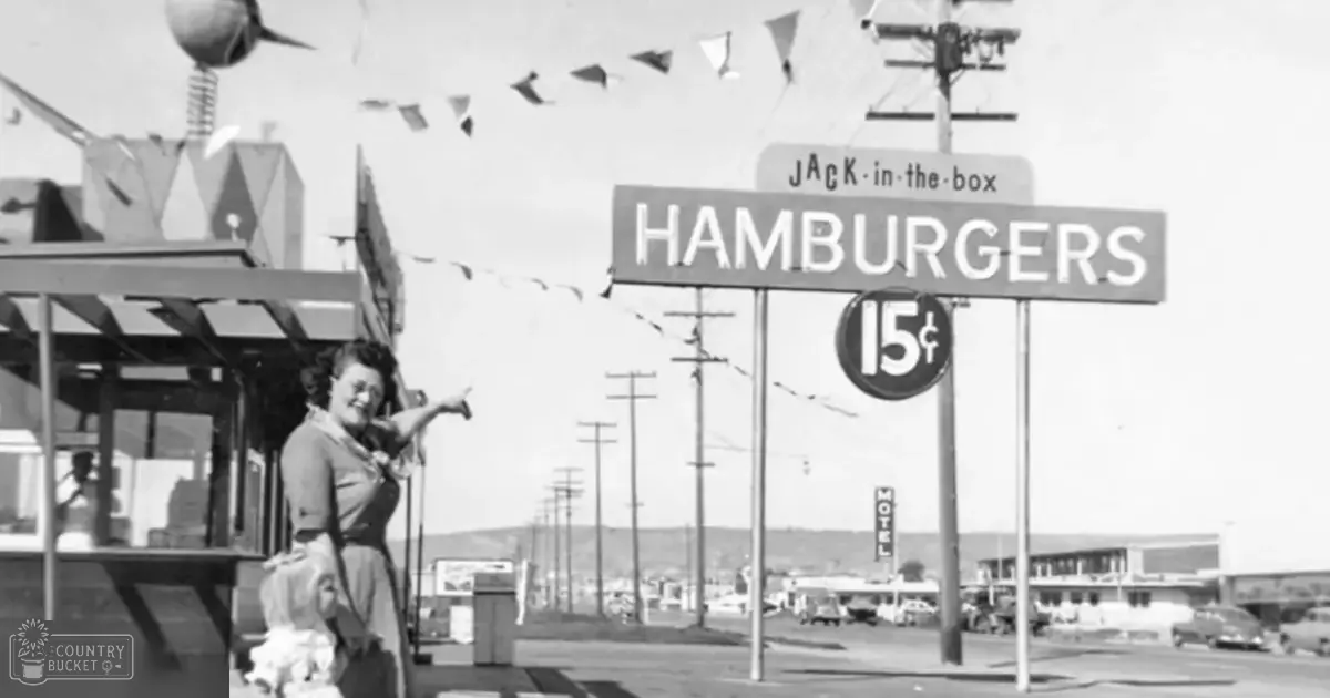 A woman in a short-sleeved dress stands on an urban street with Jack-in-the-Box signage and classic cars, while a juvenile dog with light-colored fur walks nearby. The scene suggests mid-20th century America.