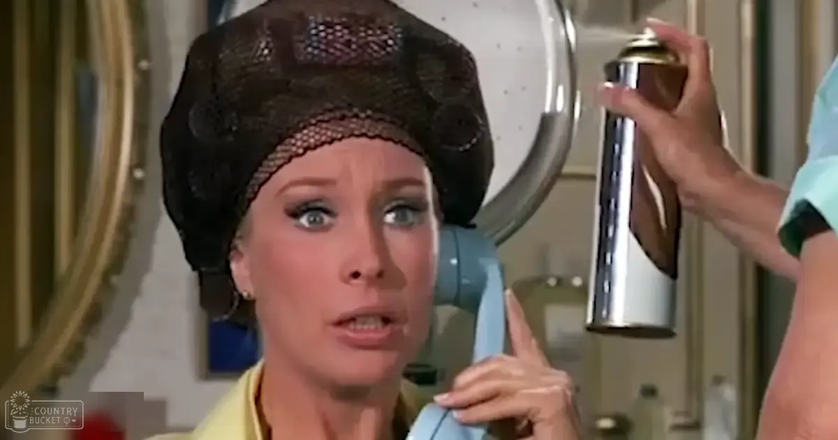 An older adult female wearing a large dark lace headpiece and yellow blouse indoors, with visible hair styling equipment and neutral-colored walls.
