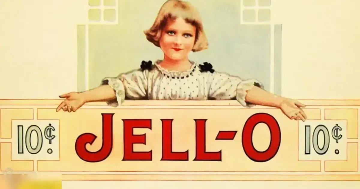 A young adult female models a white blouse with black polka dots and ruffled details in an indoor setting, wearing red JELL-O sign.