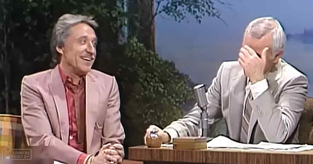 A talk show host and another person are seated at a desk with microphones, wearing formal attire. The setting is an indoor television studio with a backdrop of greenery suggesting spring or summer.