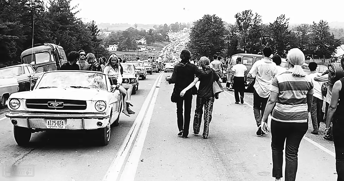 A gathering at a rural road with vintage cars and retro fashion indicative of the 1960s era.