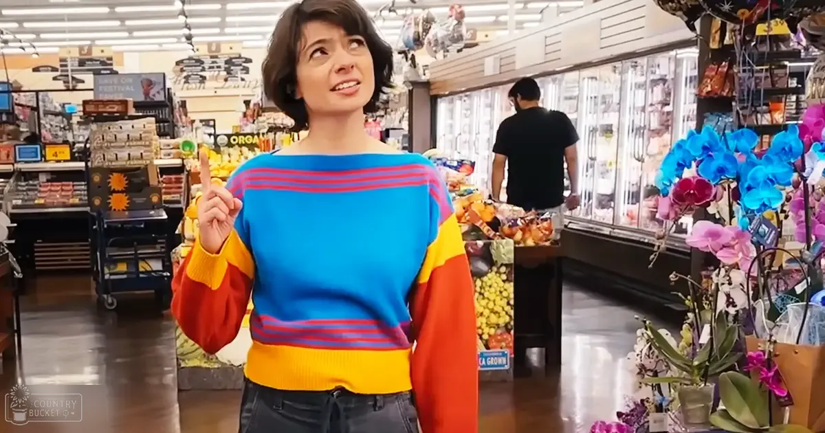 A young adult female in a grocery store, wearing a colorful sweater with horizontal stripes of blue, red, yellow, and orange, surrounded by various grocery items and produce.