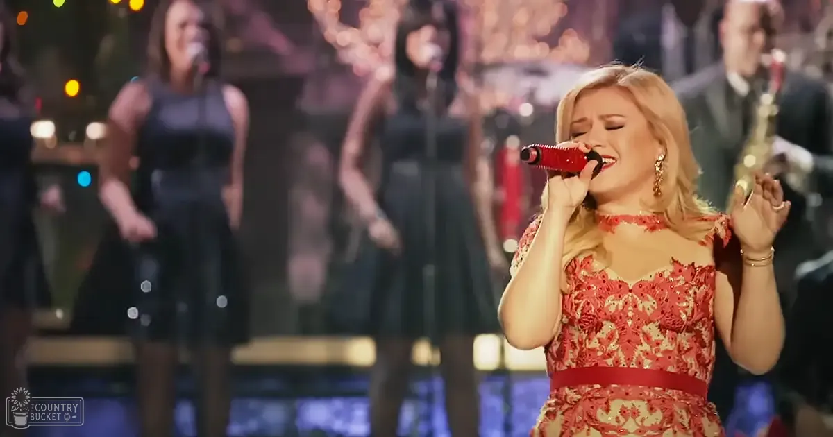 An adult female singer in a red floral-patterned dress performs on stage at an indoor concert venue with another woman in a black dress in the background.