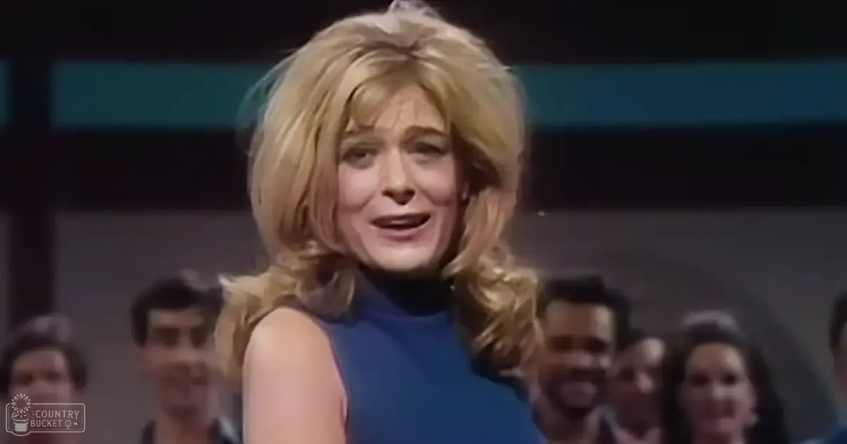 A woman with short, wavy blonde hair wearing a sleeveless blue dress at an indoor event or performance.