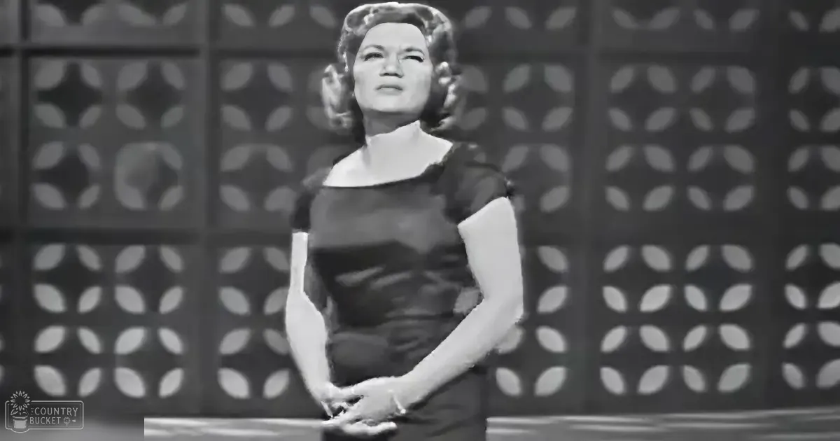 A woman wearing a form-fitting, dark-colored dress with short sleeves stands indoors against a patterned wall.