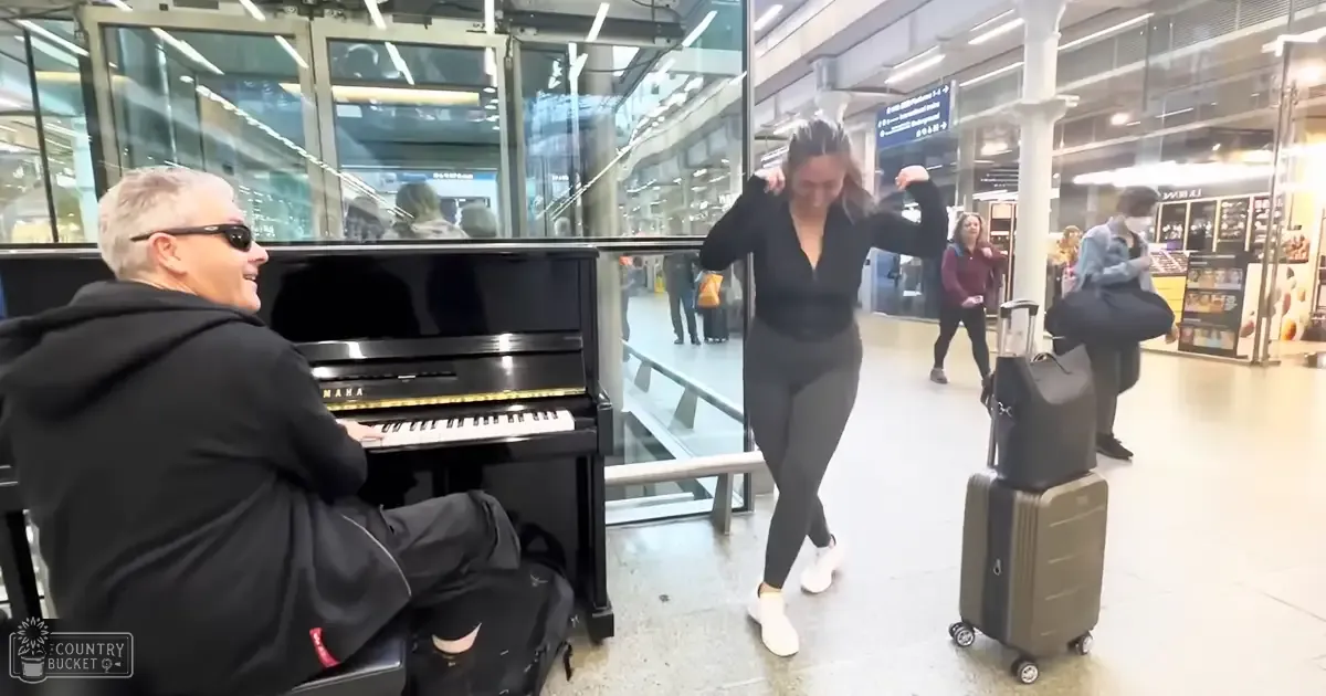 An adult male singer wearing sunglasses and a black hoodie plays the piano in an airport terminal with various people walking around. The setting is brightly lit, suggesting daytime.