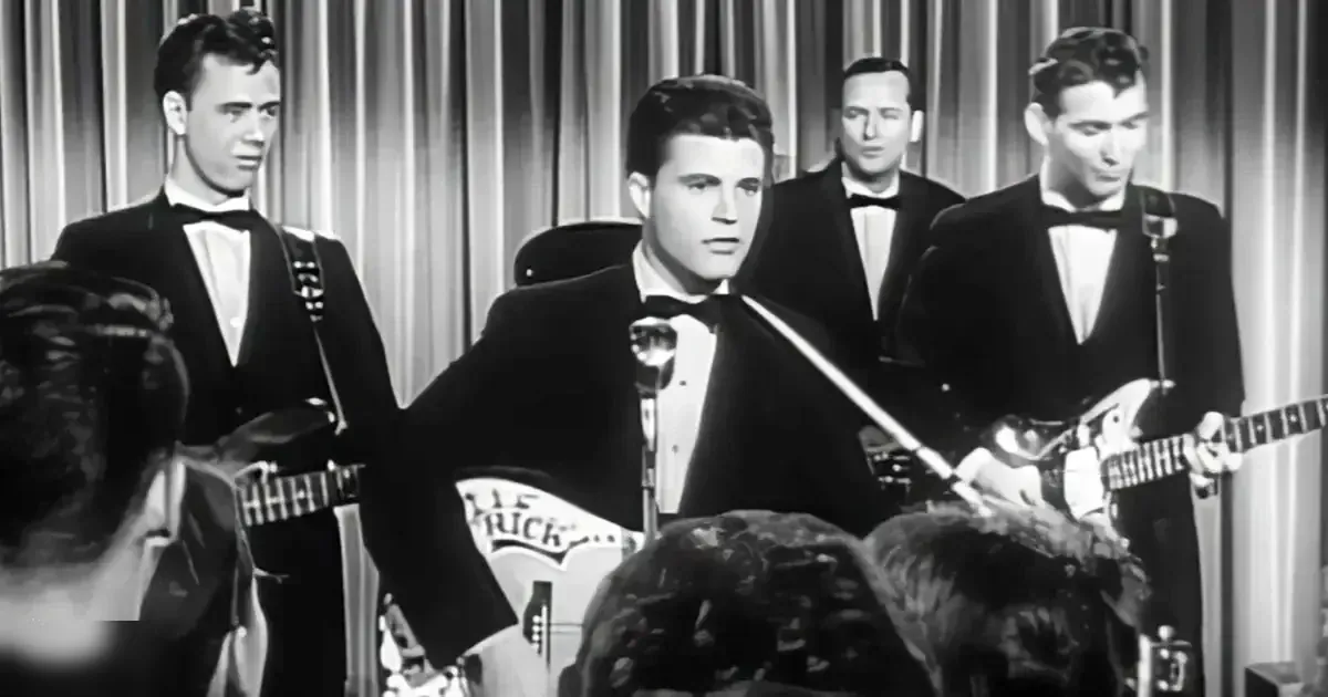 Young adult male performer in a black tuxedo with long-sleeved shirt and bow tie, surrounded by other performers in dark suits at what appears to be a concert or stage performance setting.