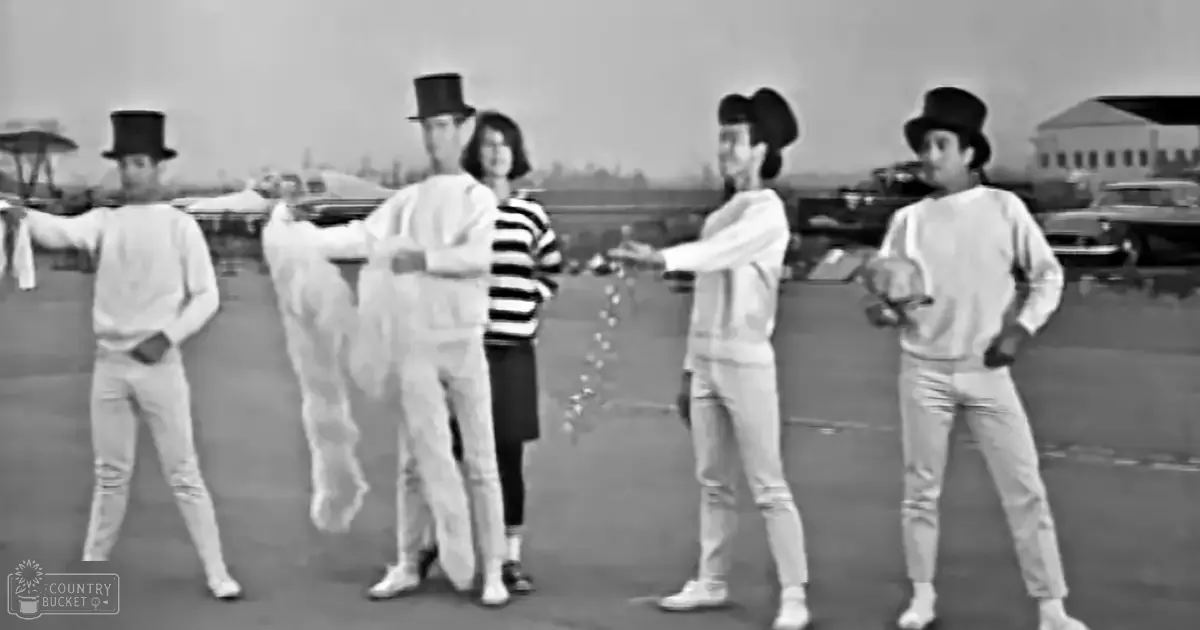 A group of young adult males in white long-sleeved shirts and light-colored pants with black top hats performing or rehearsing in a parking lot under late afternoon or early evening lighting, surrounded by parked cars.