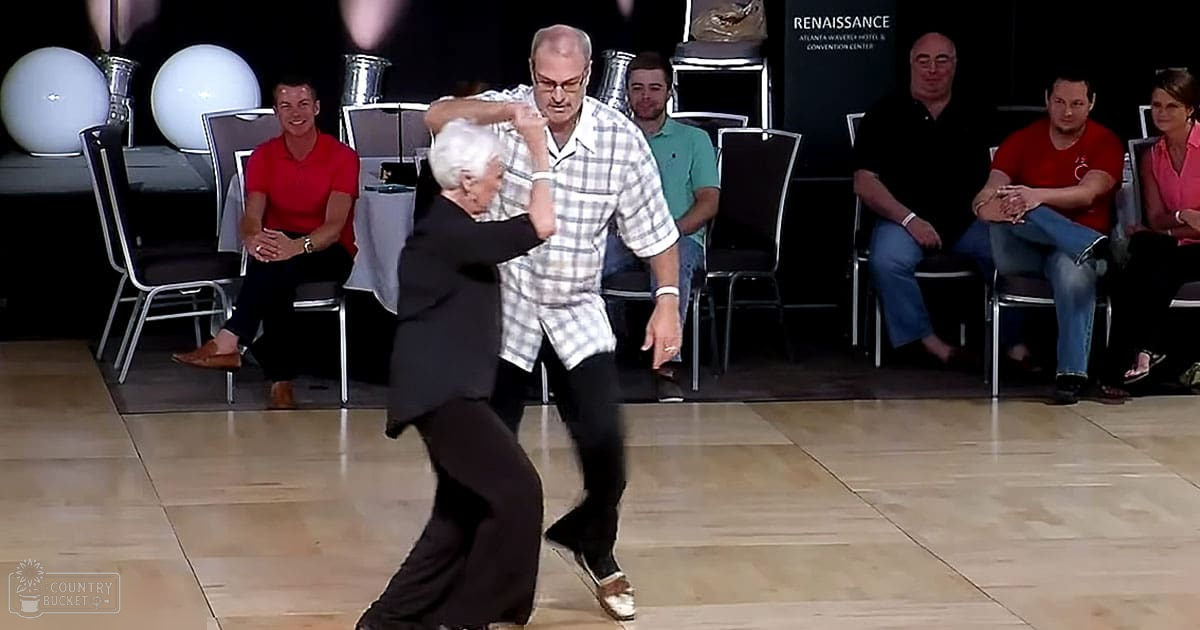 A dance duo performs at an event indoors with seated spectators; male dancer middle-aged adult wearing glasses, plaid shirt, dark pants, white shoes; female dancer older adult in black blazer, dark pants, gold shoes. Spectators include a young adult male in red t-shirt and dark pants, a middle-aged male in green polo shirt and jeans, an older male with glasses in black shirt and jeans, and an older female in pink top and dark pants seated on the far right; round white decorative objects and 'Renaissance Atlanta World Hotel & Convention Center' sign visible. The setting appears to be indoors at night or evening due to artificial lighting and darkened background.