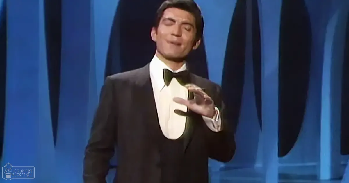 An adult male performer in a black tuxedo with white dress shirt and green bow tie, set against blue curtains typical of a theater or studio.