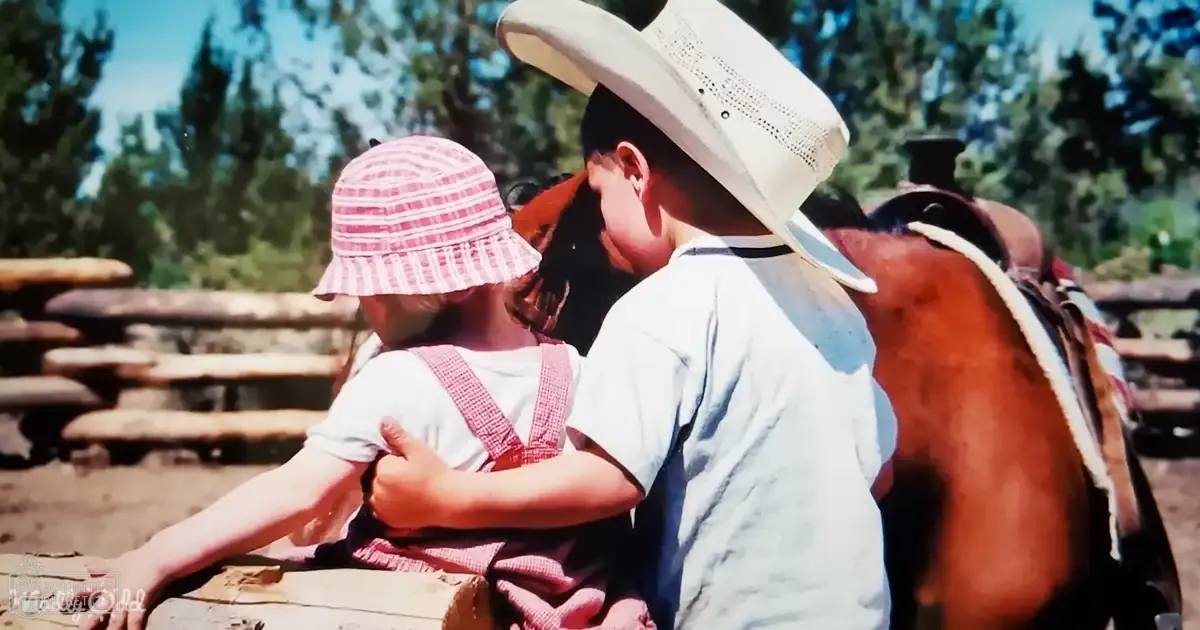 A toddler girl wearing white with red suspenders and a pink bucket hat sits on a wooden bench, accompanied by an adult male in a light shirt and wide-brimmed hat. A brown horse stands nearby in a lush green park setting during what appears to be summer or spring.
