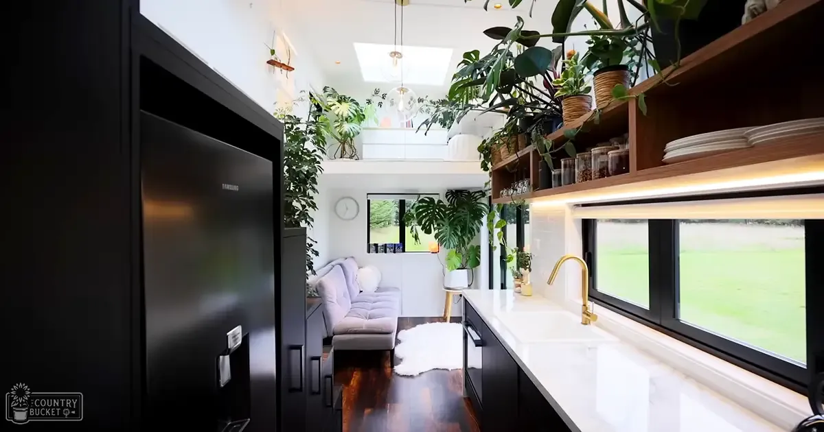 A modern kitchen with an open-plan living area in a home setting with stainless steel refrigerator, white countertop, wooden shelves with plants and dishes, gray sofa with cushions near clock, potted plants, small wooden stool, skylight allowing natural light into the space.