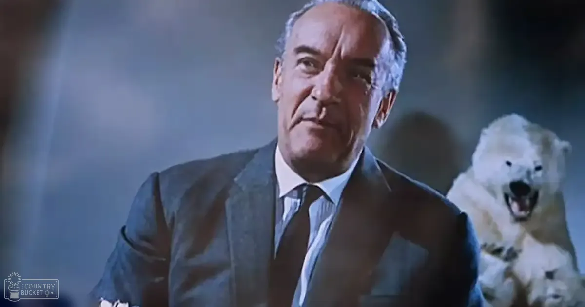 An adult male in a dark patterned suit jacket and light dress shirt stands indoors, possibly urban, with even soft lighting and muted blue-gray tones; polar bear in the background.