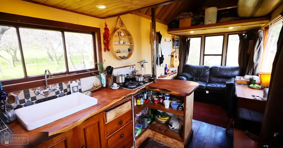 A cozy, rustic kitchen interior with a wooden countertop, checkered backsplash, black leather couch, guitar hanging on the wall, and greenery visible through a window in a cabin or small home during daytime.