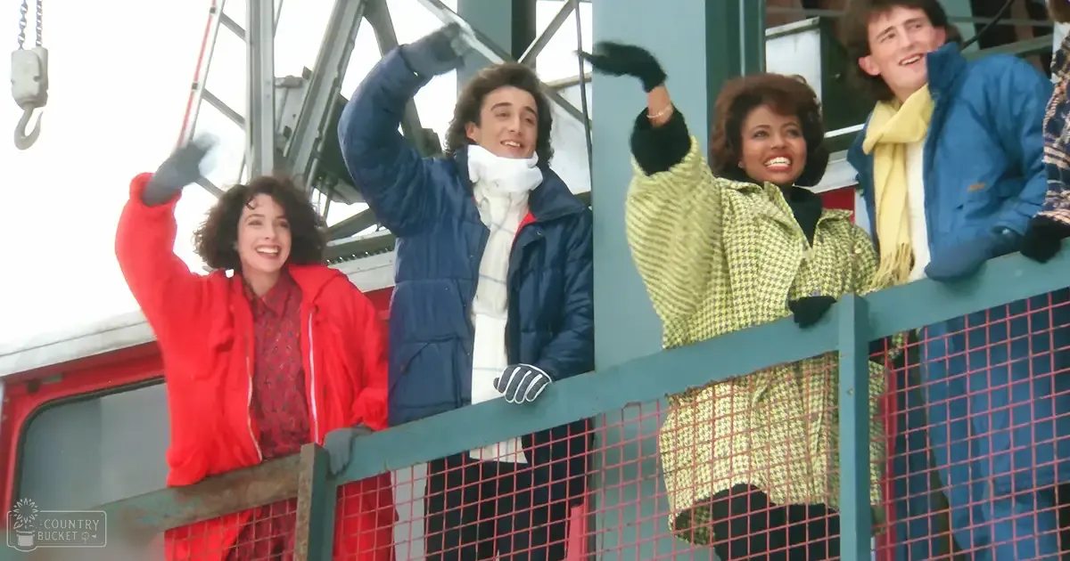 A group of friends enjoys an outdoor outing in a cold, winter setting with heavy jackets and gloves, surrounded by metal structures and cranes at what appears to be an industrial site. Natural daylight suggests late afternoon.