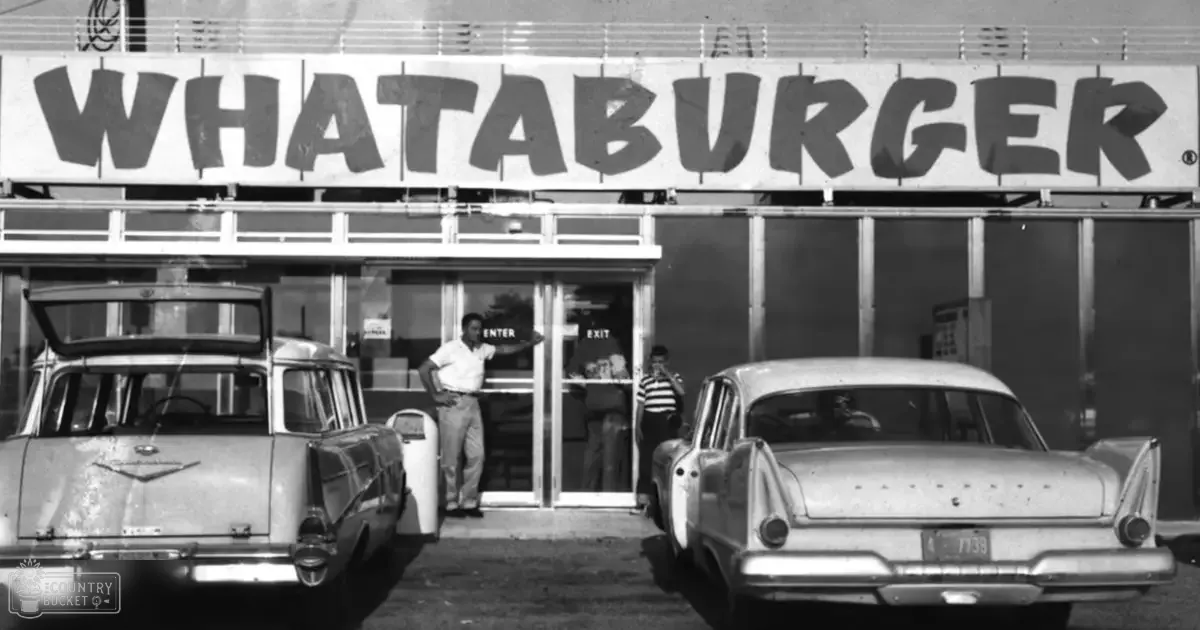 A Whataburger restaurant with two vintage cars parked outside; an adult male and female stand near the entrance, in a black-and-white daytime image.
